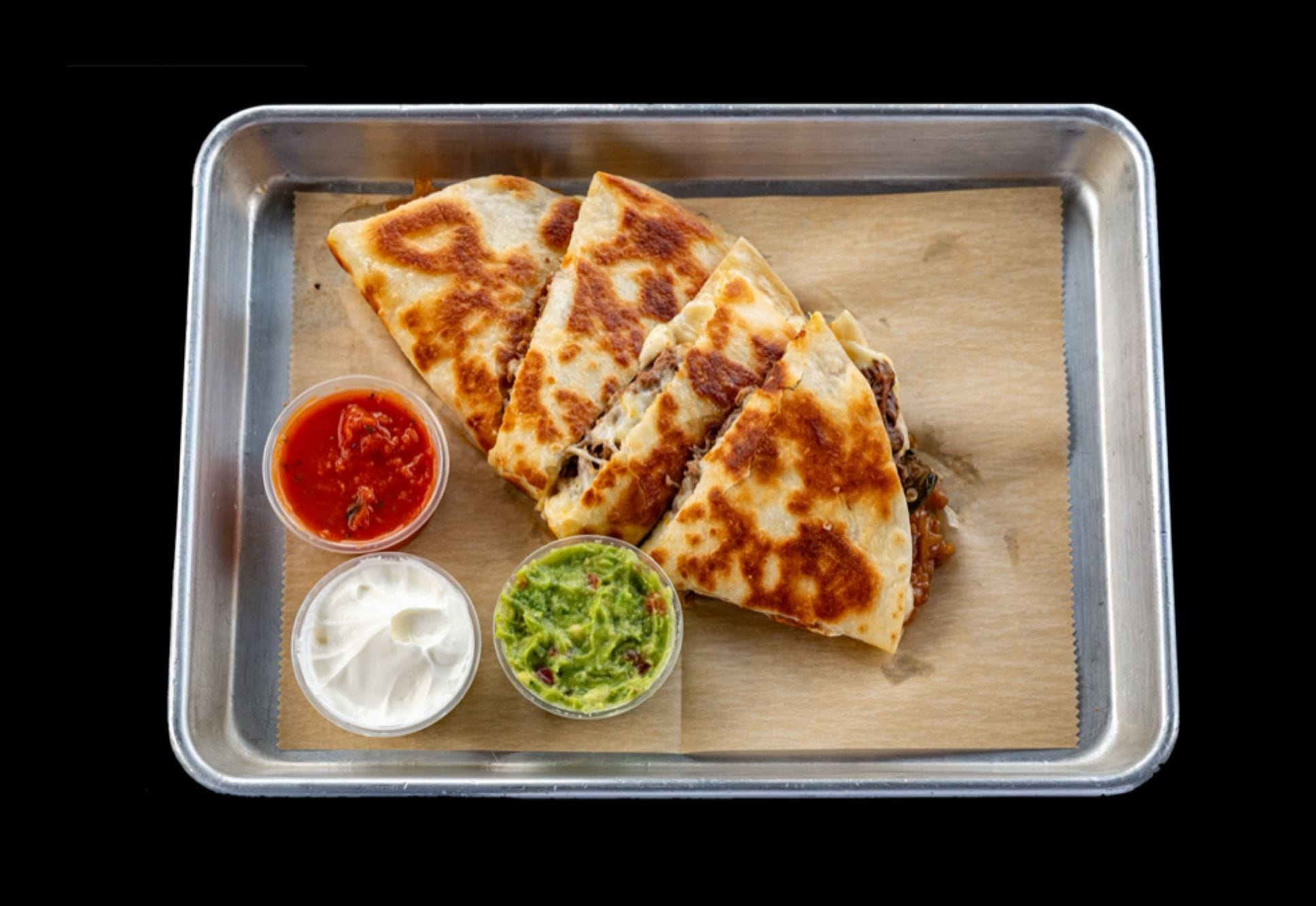 Golden-brown quesadilla served on a plate with salsa, guacamole, and sour cream garnishes. Cut into wedges and filled with melted cheese, grilled onions, jalapenos, and choice of protein including steak, chicken, chorizo, al pastor, barbacoa, tinga, or carnitas.